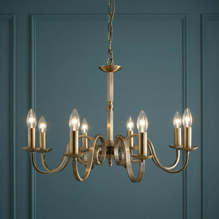 Richmond 8-light antique brass chandelier with elegant scroll arms and candle bulbs against a teal panelled wall.