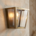 Antique brass box wall light with glass panels and a glowing bulb mounted on a textured beige wall.