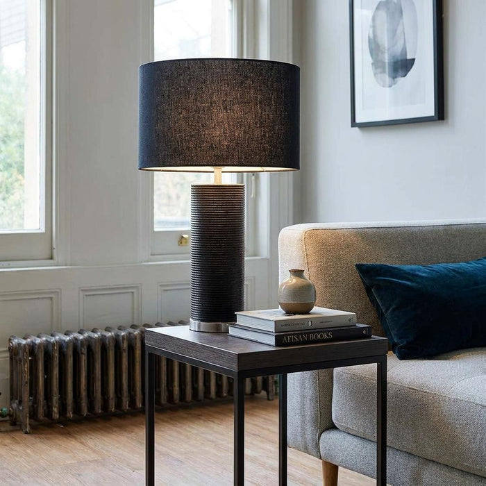 Black textured table lamp with a dark shade on a side table next to a sofa in a bright living room.