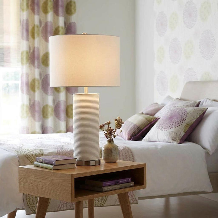A Ripple 1 light white table lamp with a ribbed ceramic base and drum shade on a wooden bedside table in a bedroom.
