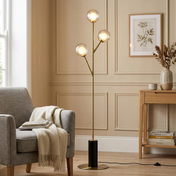 Riviera Brass Art Deco Floor Lamp with three ribbed glass globes, placed beside a grey armchair in a living room.