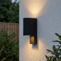 Modern black up and down wall light mounted on a textured white exterior wall, casting warm pools of light.