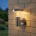 Illuminated Roko brushed stainless steel outdoor wall light with clear glass shade mounted on a brick wall.