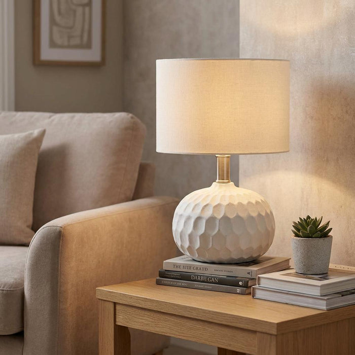 White ceramic table lamp with a textured base and beige shade on a wooden side table in a neutral living room setting.