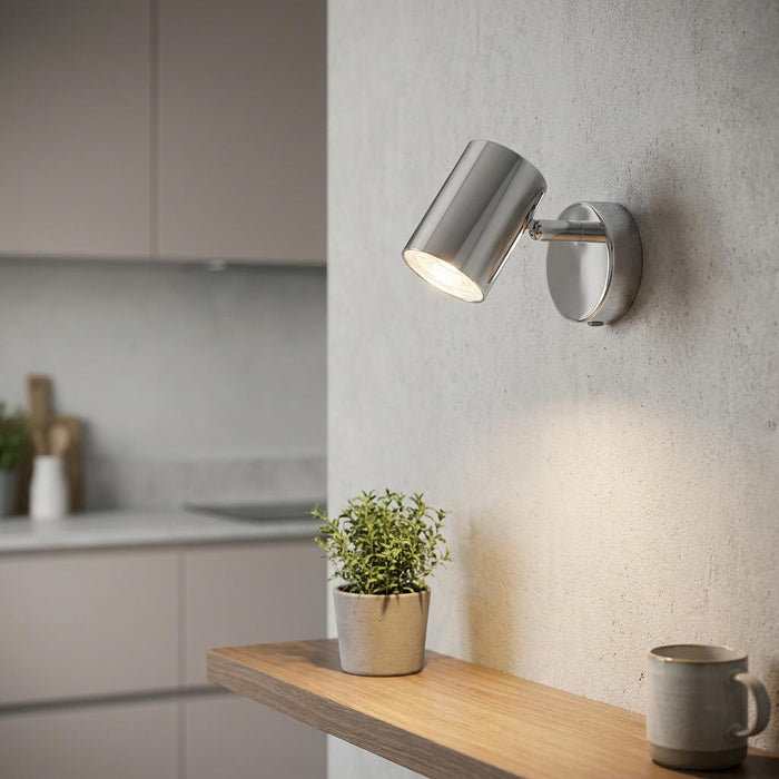 A modern wall-mounted spotlight with a brushed silver finish, shown lit on a textured wall above a wooden shelf in a kitchen setting.