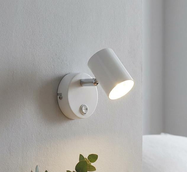 White cylinder adjustable wall spotlight illuminating a bedside table with a plant and books.