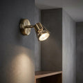 Romore antique brass single spotlight mounted on a textured grey wall, casting a warm glow.