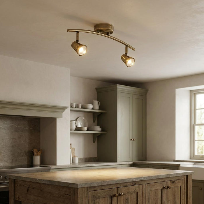 Antique brass twin kitchen spotlight with curved bar and adjustable heads illuminating a rustic wooden island.