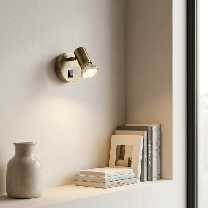 Antique brass switched spotlight mounted on a wall, illuminating books on a minimalist shelf.