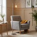 A slim Rubens Chrome Floor Reading Lamp with an exposed bulb stands next to a grey armchair in a cozy living room.