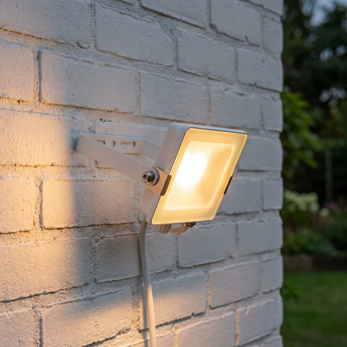 White square LED flood light illuminated and mounted on an exterior white brick wall.