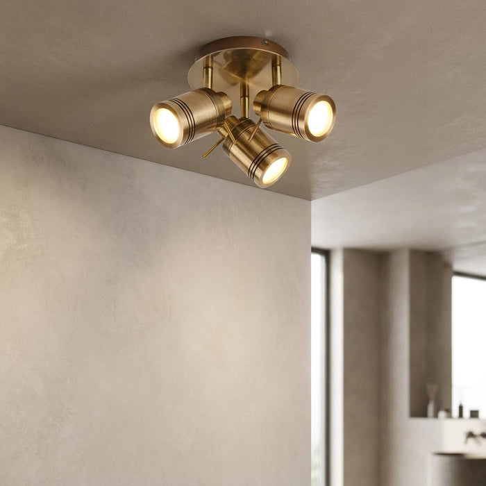 A modern brass flush mount ceiling light with three adjustable cylindrical spotlights illuminating a neutral room.