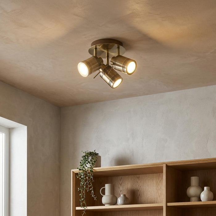 Samson three-light brass bathroom adjustable flush spotlight glowing on a textured ceiling above wooden shelving.