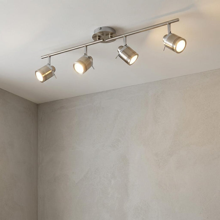 Adjustable four-light silver split-bar spotlight fixture mounted on a white ceiling above a grey textured wall.