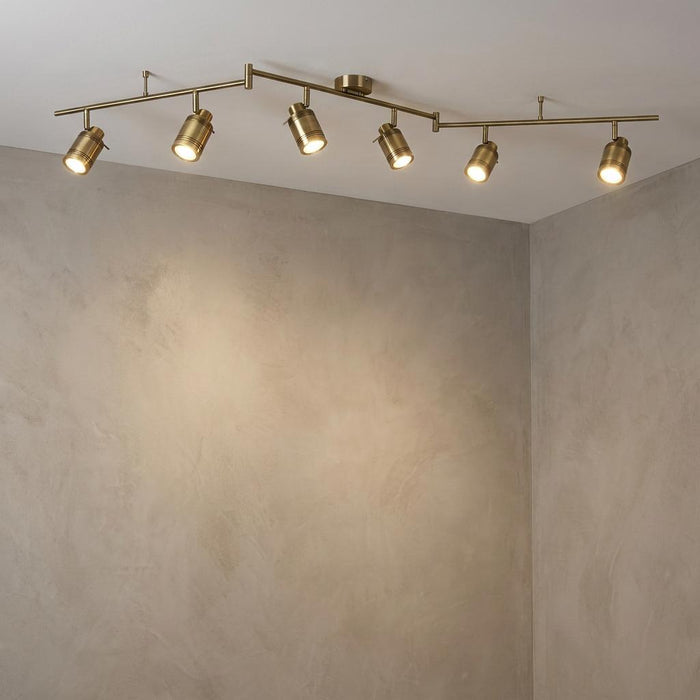 A brass six-light adjustable split-bar spotlight mounted on a ceiling, illuminating a grey textured wall.