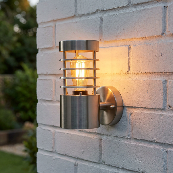 Brushed steel outdoor wall light with a glowing filament bulb mounted on a white brick wall.