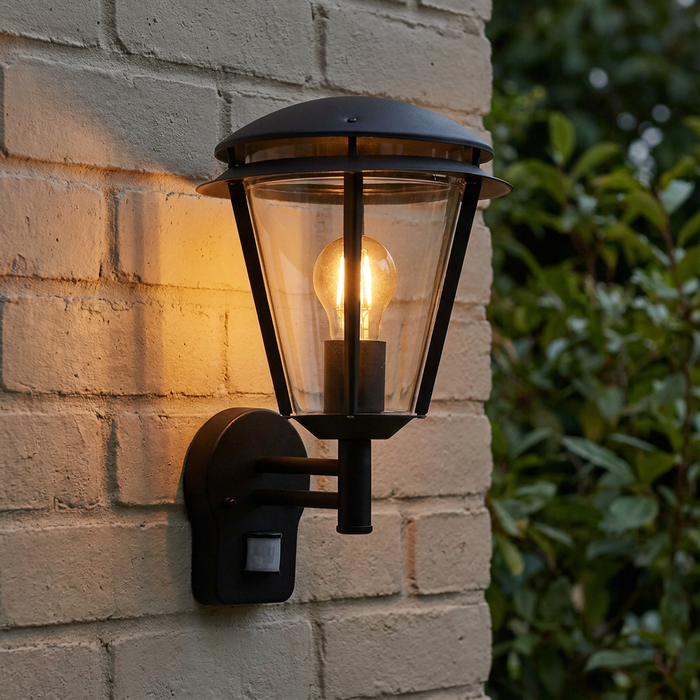 Matt black outdoor PIR wall light with a glowing filament bulb, mounted on a light brick wall.