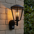 Matt black outdoor PIR wall light with a glowing filament bulb, mounted on a light brick wall.