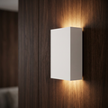 White rectangular plaster wall light on a dark wood wall, casting a warm up-and-down glow.
