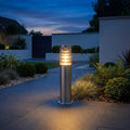 A brushed stainless steel outdoor bollard light illuminates a modern stone-paved garden path at twilight.