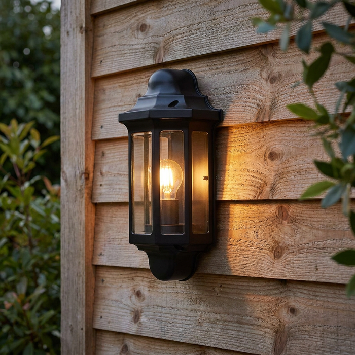Traditional satin black lantern wall light with a glowing bulb mounted on a wooden fence.