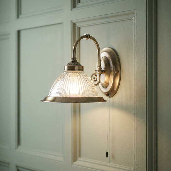 Antique brass wall light with a ribbed glass shade and pull cord mounted on a green paneled wall.