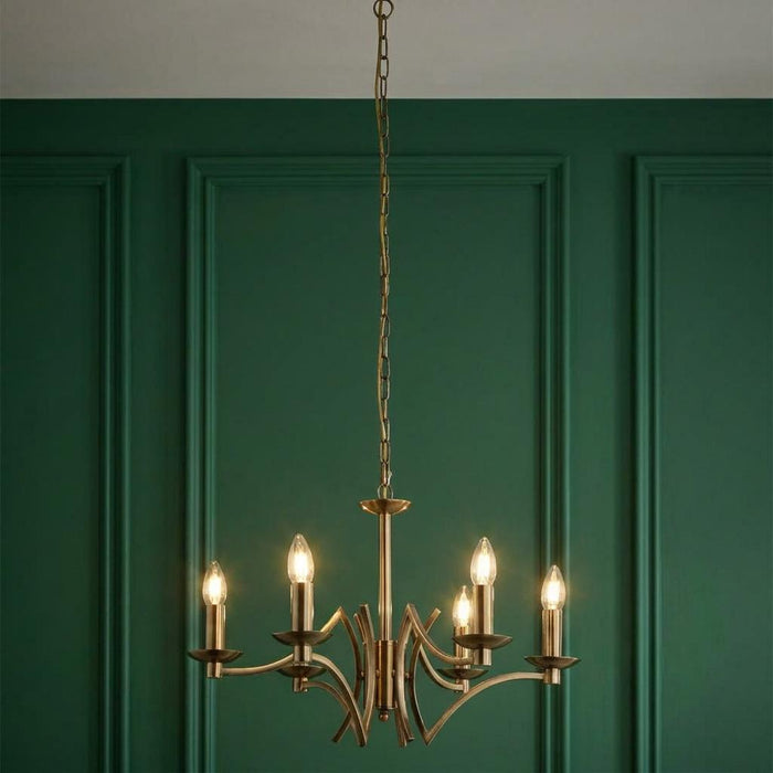 Searchlight Ascot 6-light antique brass chandelier with curved arms against a green paneled wall.