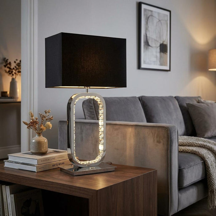Chrome table lamp with a black rectangular shade and crystal-lined oval base on a wooden side table next to a sofa.