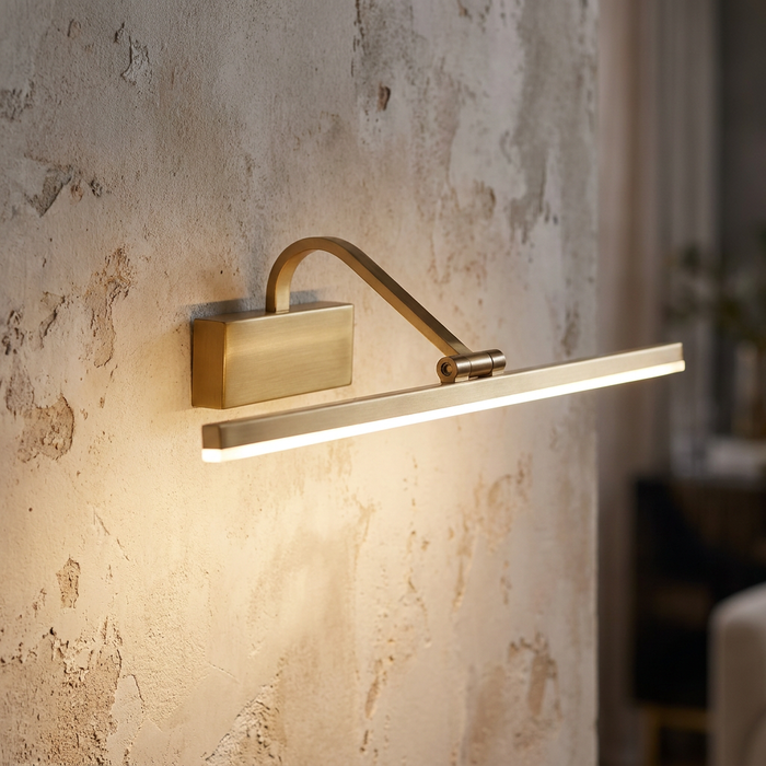 A minimalist bronze LED picture light mounted on a textured plaster wall, casting a warm downward glow.