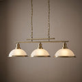 A brass three-light bar pendant ceiling fixture with white frosted glass dome shades, illuminated against a neutral wall.