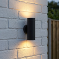 Black cylindrical outdoor wall light emitting warm up and down glows on a white brick wall.