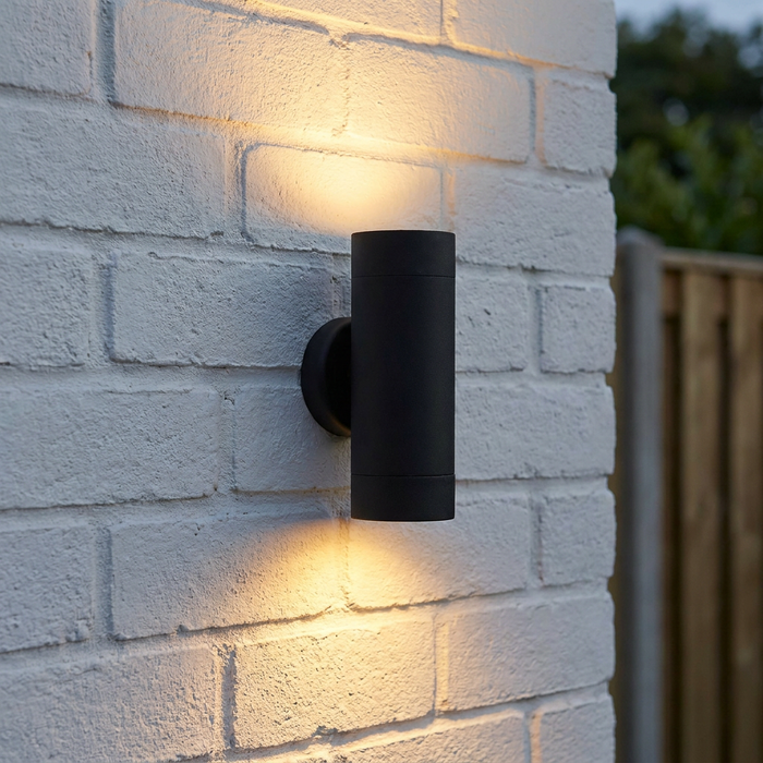 Black cylindrical outdoor wall light emitting warm up and down glows on a white brick wall.