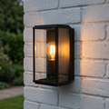 A black rectangular box wall lantern with a warm filament bulb mounted on a white brick wall.