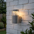 Stainless steel cylindrical LED outdoor wall light mounted on a white brick wall, casting a warm glow.