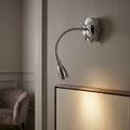 A polished chrome LED reading light with a flexible neck, mounted on a neutral wall above a framed piece.