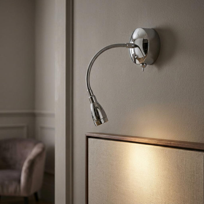 A polished chrome LED reading light with a flexible neck, mounted on a neutral wall above a framed piece.