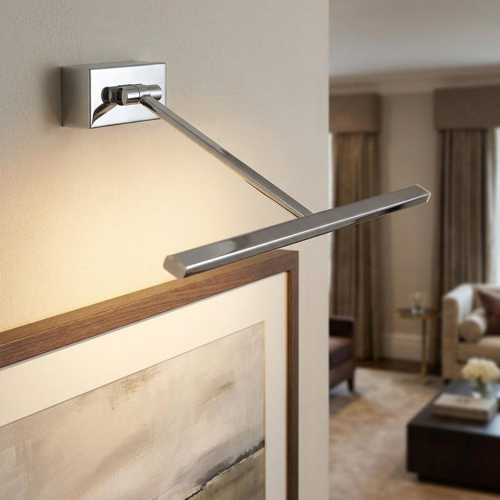 Modern chrome LED picture light mounted on a wall illuminating a framed artwork in a living room setting.