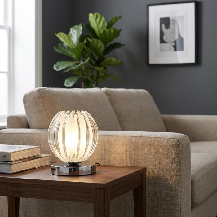 Chrome and ribbed clear acrylic table lamp glowing on a wooden side table in a modern living room.