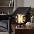Spherical table lamp with smokey acrylic ribs glowing on a wooden side table next to a dark grey armchair.