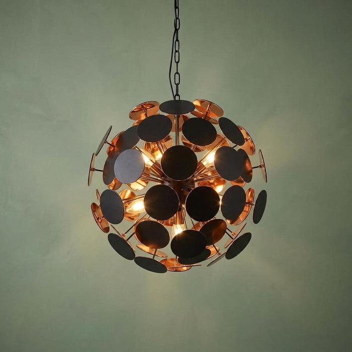 Modern spherical pendant light with matte black and gold discs illuminating against a sage green wall.