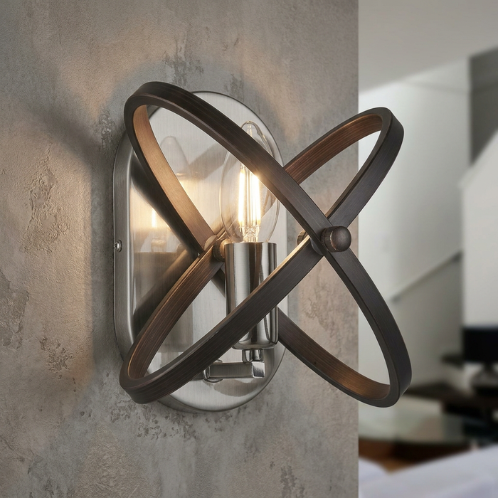 Modern pewter wall light with interlocking bronze hoops surrounding a glowing filament bulb on a textured grey wall.