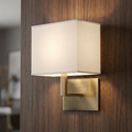 Brass wall light with a white rectangular shade mounted on a wooden wall, emitting a warm glow.