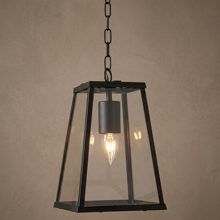 Black tapered lantern pendant light with clear glass panels and a lit candle bulb, hanging from a black chain.