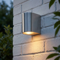Brushed silver cylindrical wall light casting a warm downward glow on a white brick exterior wall.