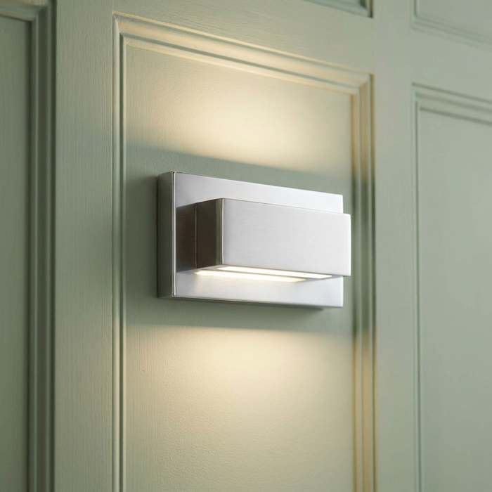 Modern rectangular chrome up and down wall light glowing on a sage green paneled wall.
