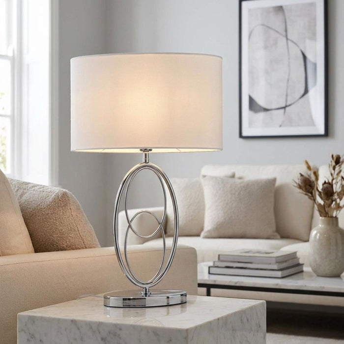 Chrome table lamp with interlocking oval base and white drum shade on a marble table in a living room.