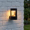 Black cylindrical outdoor wall light with warm LED illumination mounted on a white brick wall.