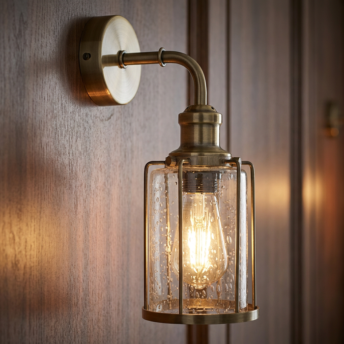 Industrial brass wall light with a seeded glass cylinder and caged frame mounted on wood paneling.