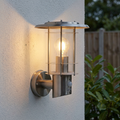 Stainless steel outdoor PIR wall light with a glowing bulb mounted on a white textured wall.