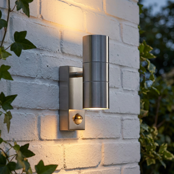 Illuminated stainless steel up/down LED outdoor PIR wall light mounted on a white brick wall.